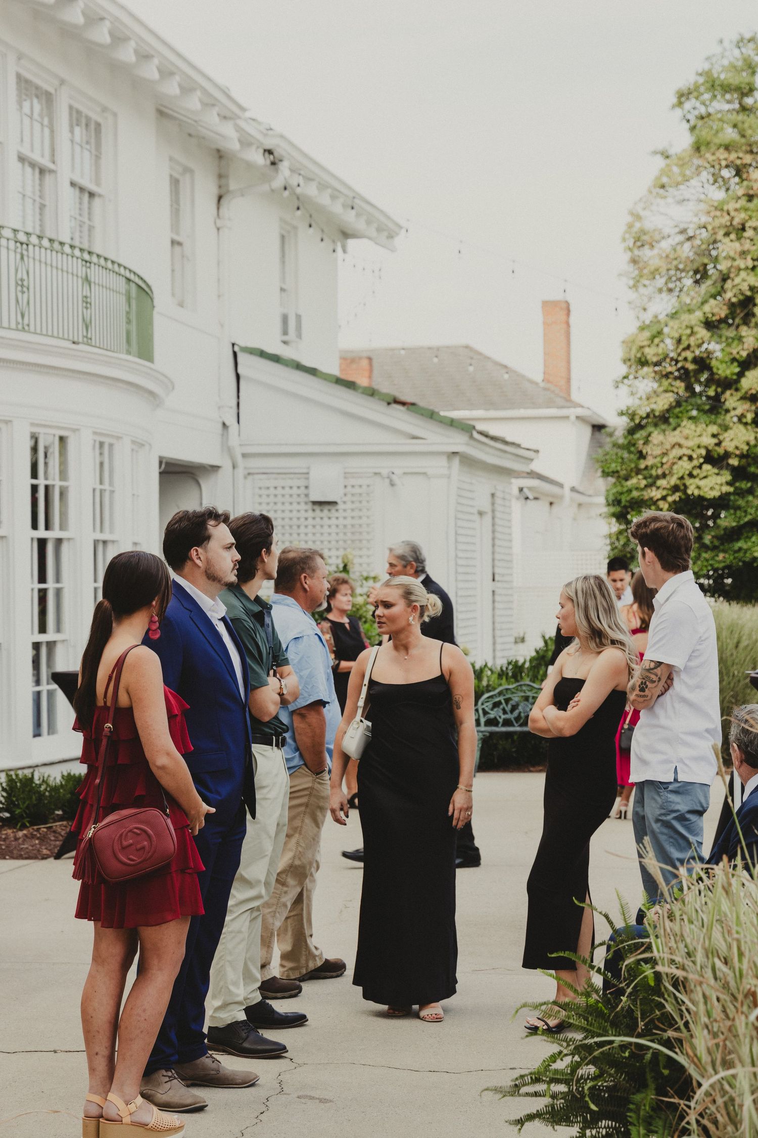 A group of friends gathered outside a white-painted building with elegant architecture and landscaping on a sunny day.