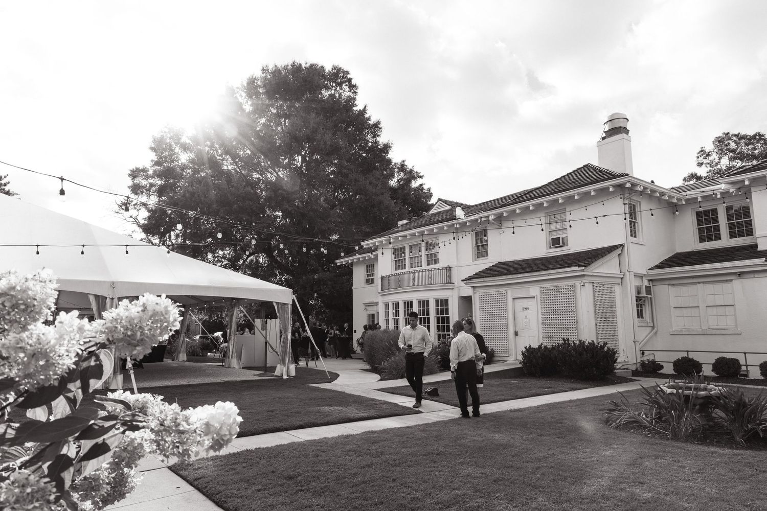 A black and white photo of a wedding venue with string lights and floral decorations on the lawn.