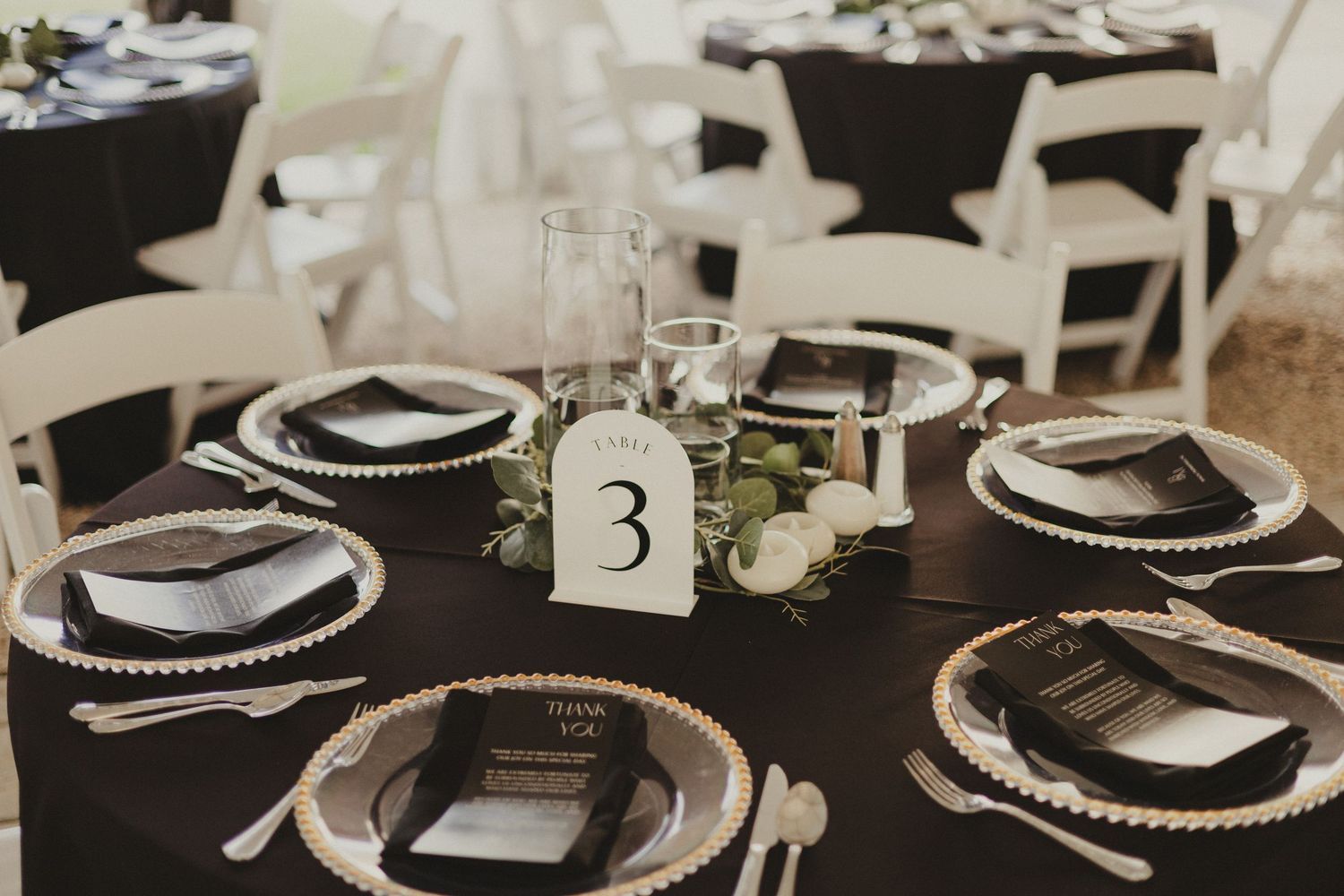 Elegant wedding table setting with black tablecloth, silver charger plates, white napkins and table number three centerpiece.