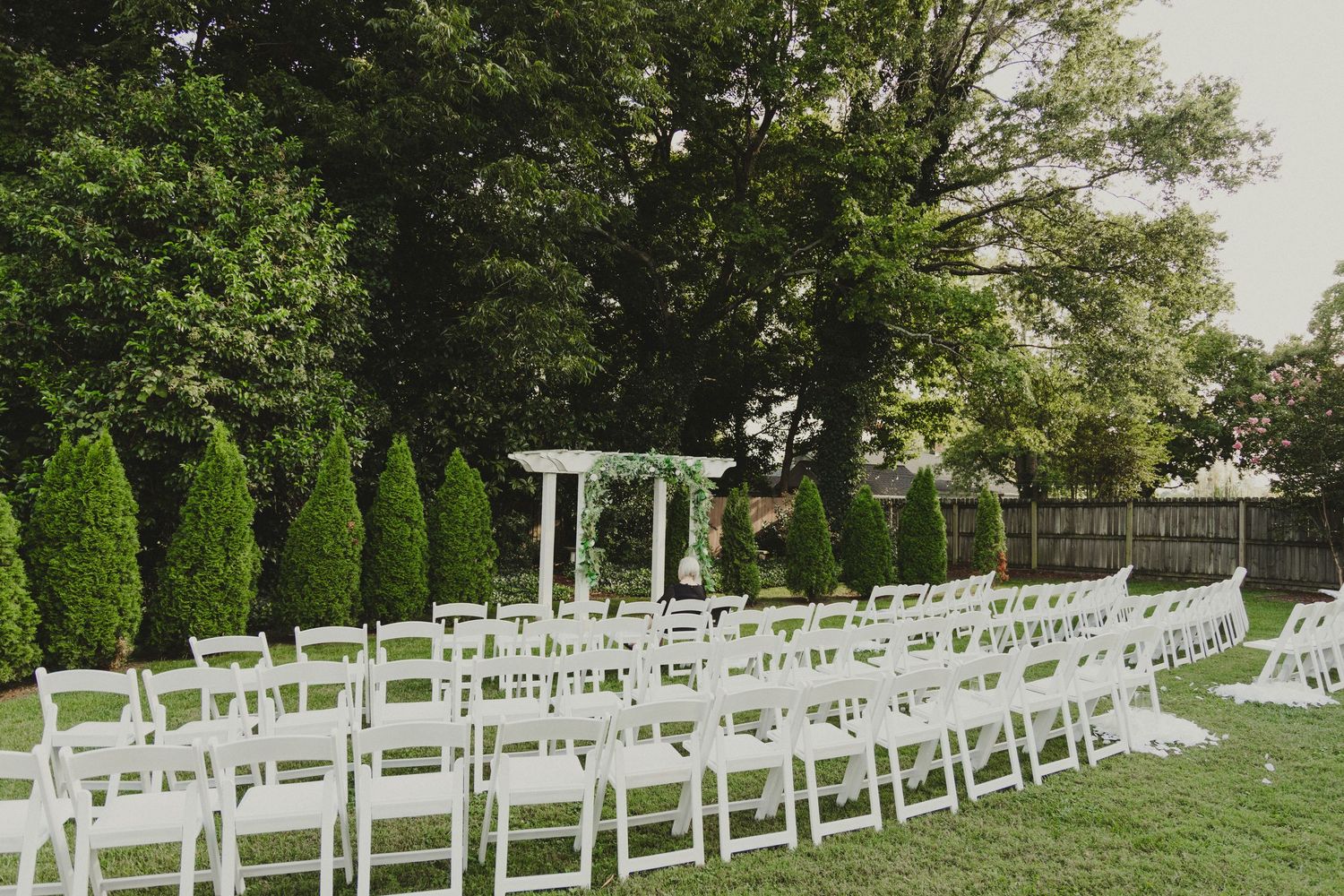 White folding chairs arranged in rows on a green lawn for an outdoor wedding ceremony with a wooden pergola and evergreens.