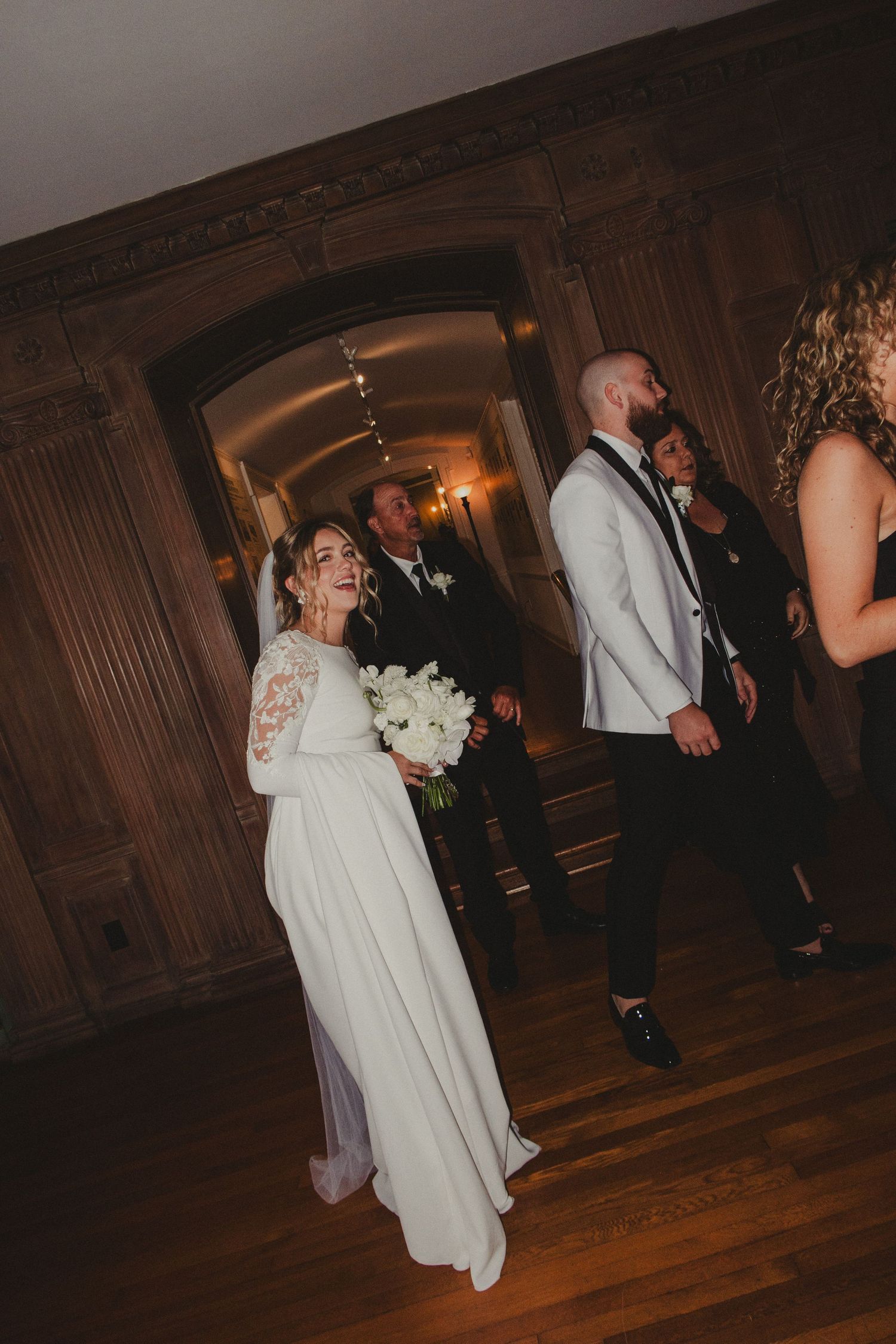 Wedding guests celebrate in an elegant wood-paneled hallway as a bride in a long-sleeved white gown holds a bouquet.