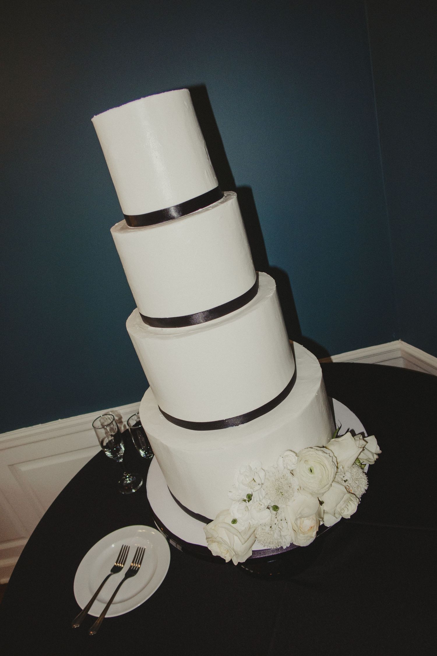 Elegant four-tier white wedding cake with brown ribbon trim and white floral decorations sits on dark table with place setting.