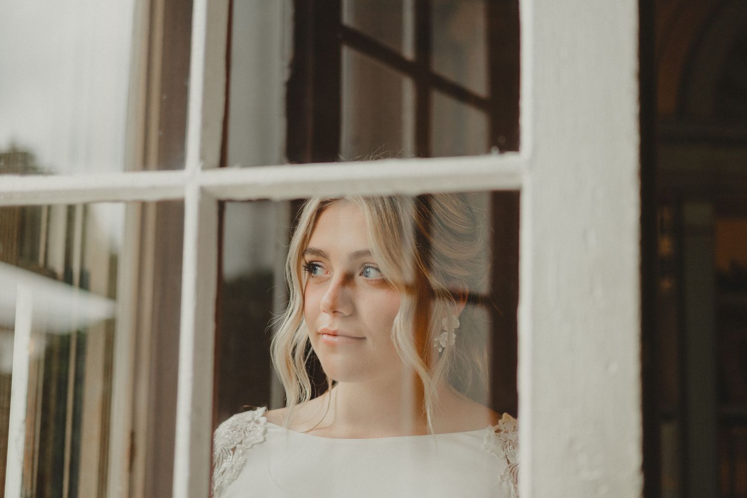 An elegant bride in a white dress stands by a window, with soft natural light illuminating her silhouette.