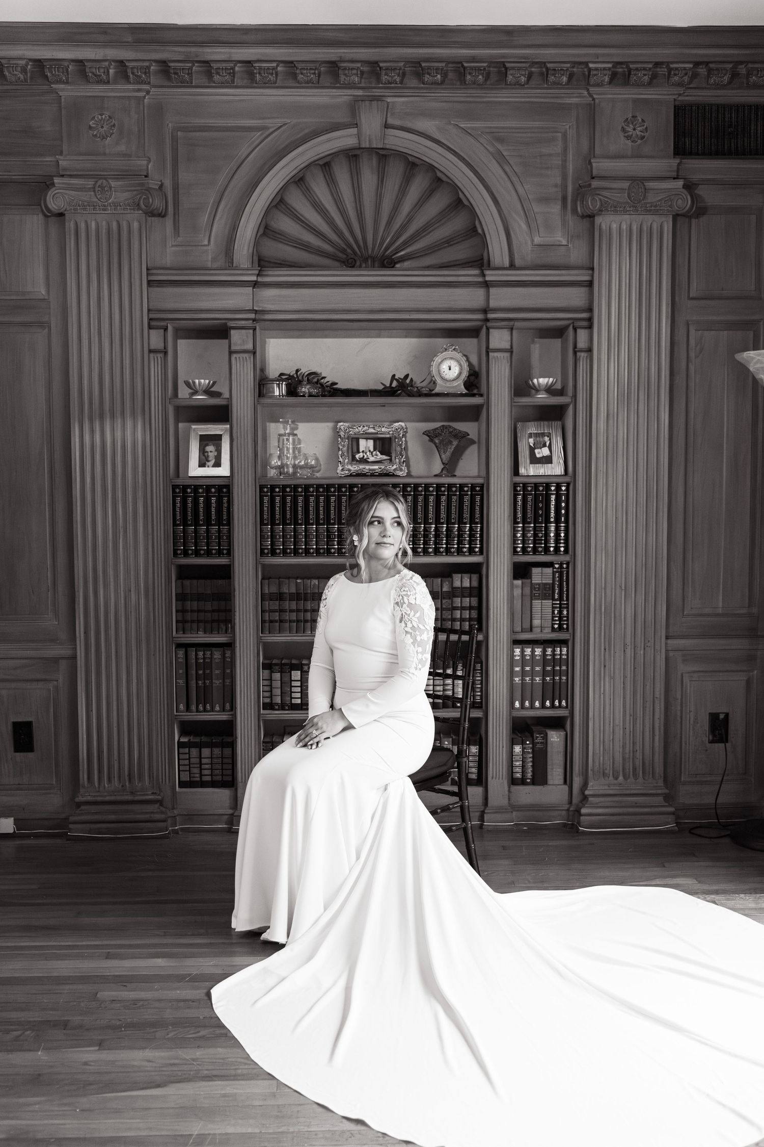 A figure in an elegant long-sleeved white wedding gown sits elegantly in front of a classic wood-paneled library with built-in shelves.