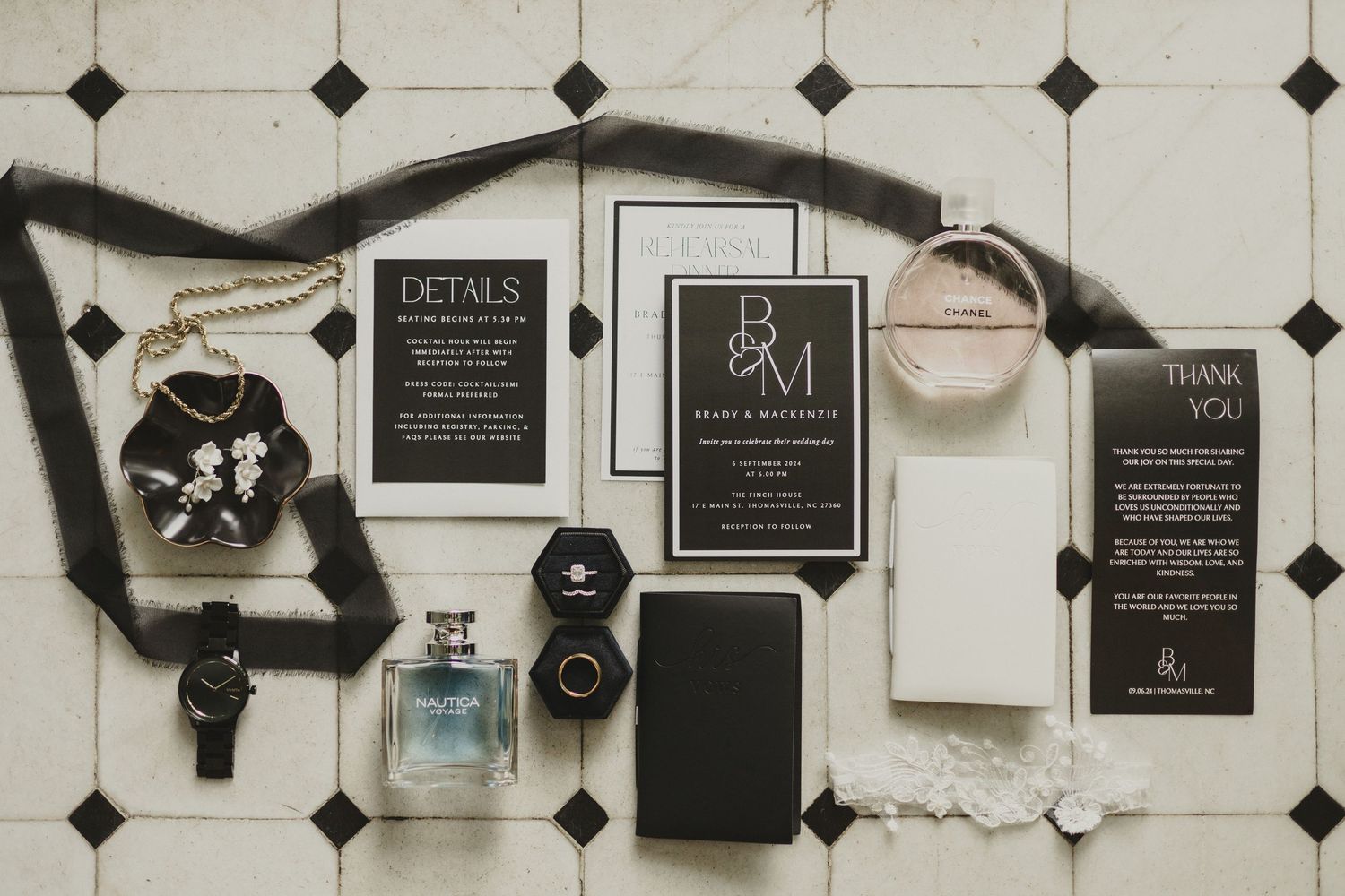 Wedding invitation suite with black and white cards, perfume bottle, and accessories laid out on geometric tile floor.