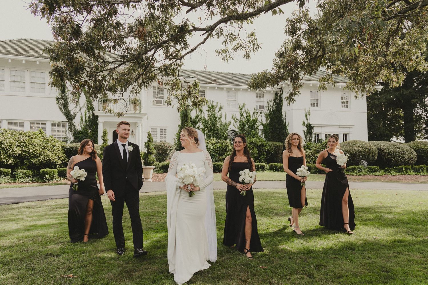 Wedding party in formal attire poses on a manicured lawn in front of a white building during an outdoor ceremony.