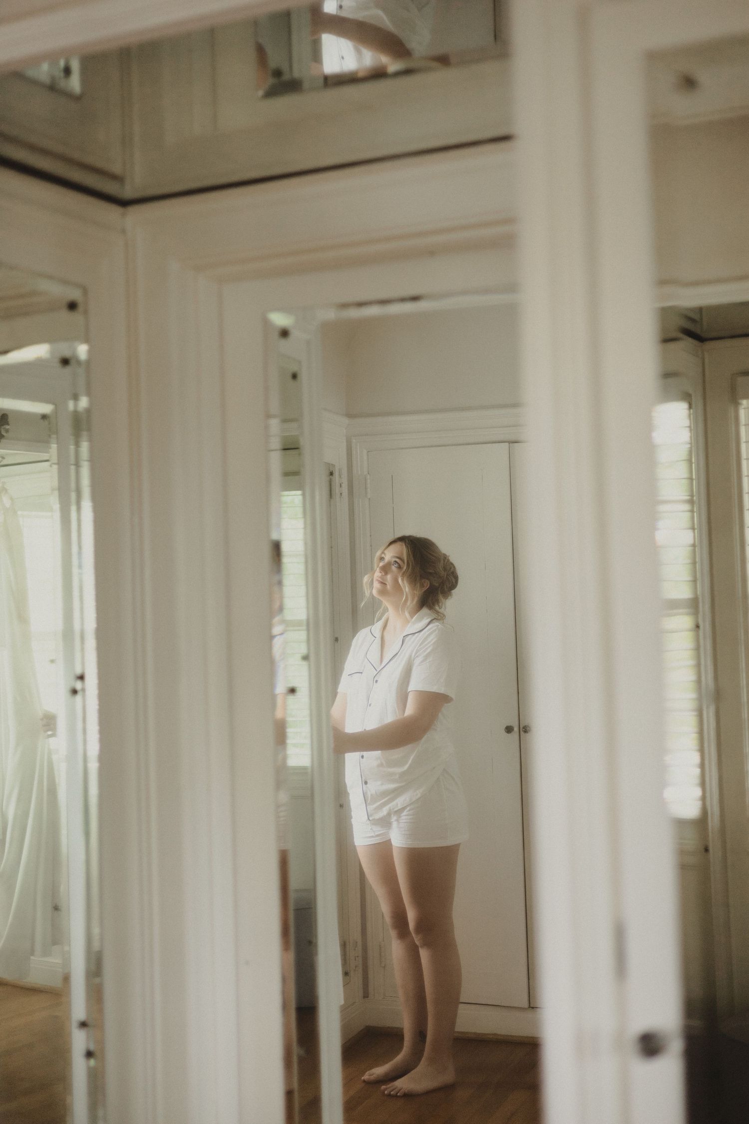 A dreamy portrait in white tones shows a figure in shorts and shirt standing between mirrors in a bright sunlit room.