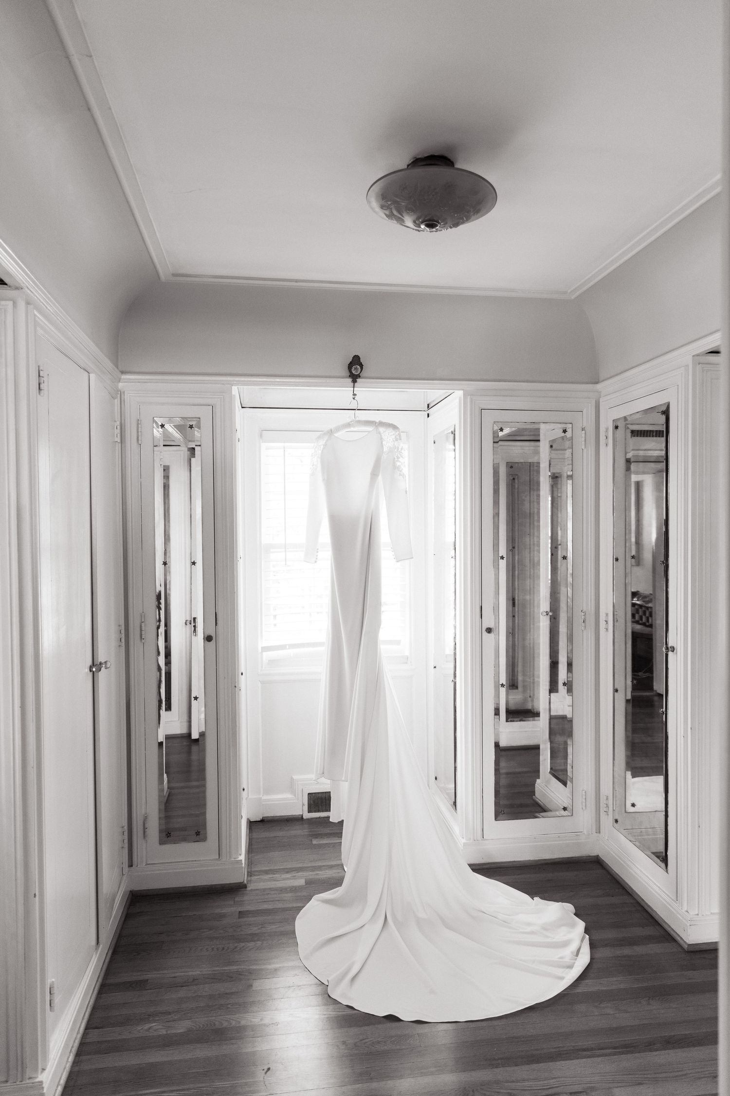 An elegant white wedding dress hangs gracefully in a bright dressing room with mirrored closet doors and hardwood floors.