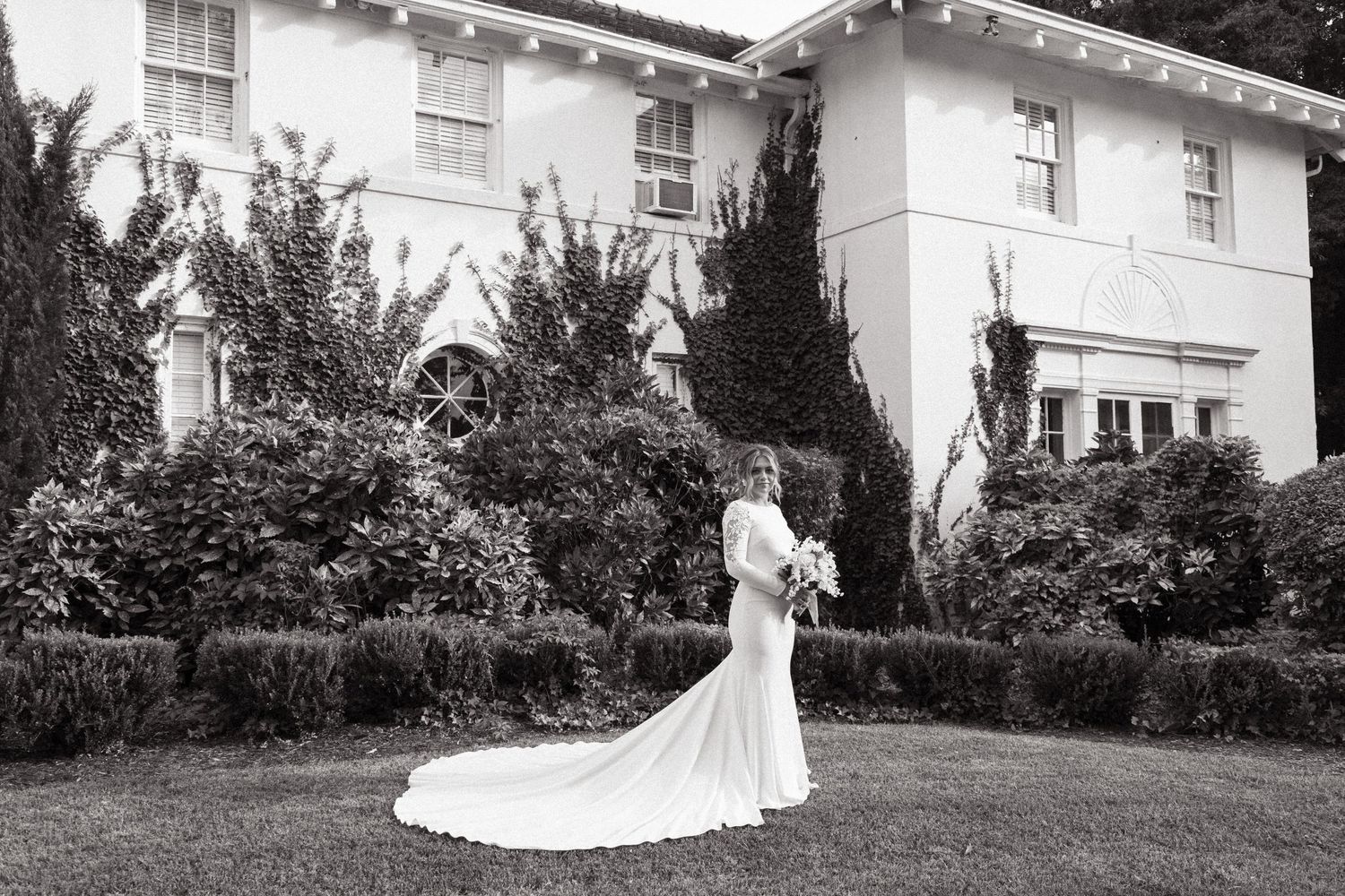 Black and white photo of a bride in a flowing white dress with long train standing in front of a stately white house.