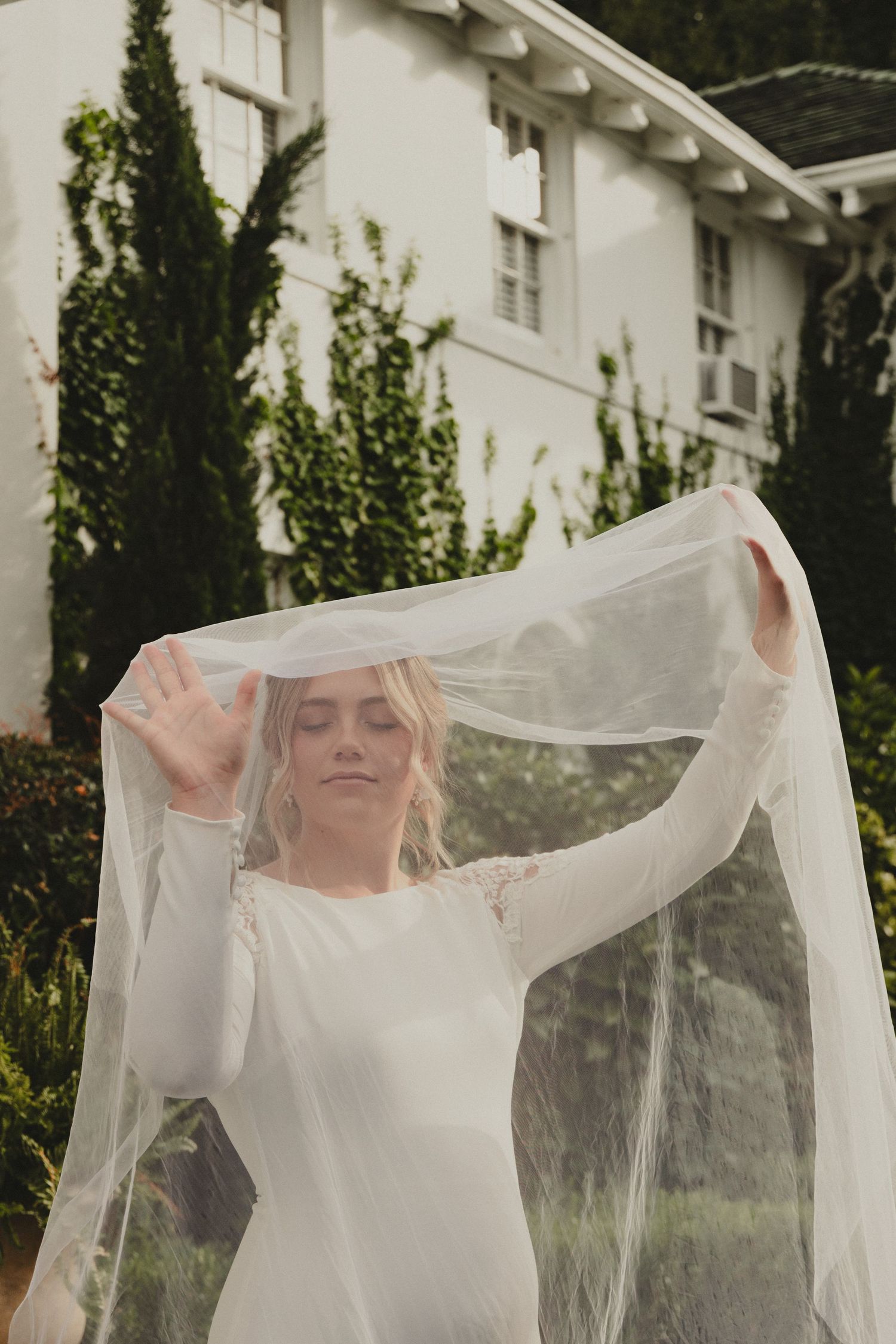 A bride in a long-sleeved white wedding dress lifts a delicate veil in front of a white building with climbing ivy.