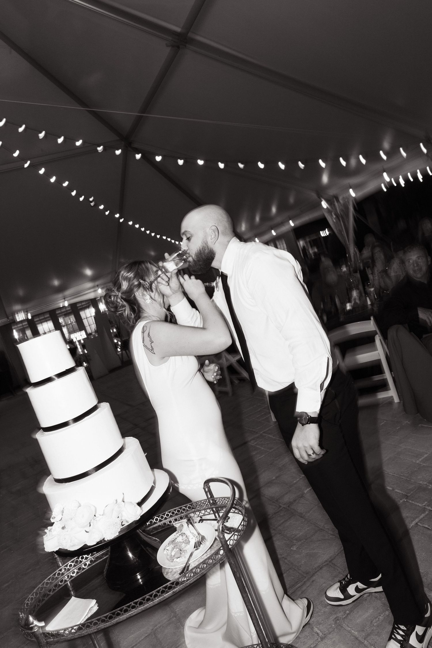 A black and white wedding photo with a couple kissing next to their tiered wedding cake under string lights.