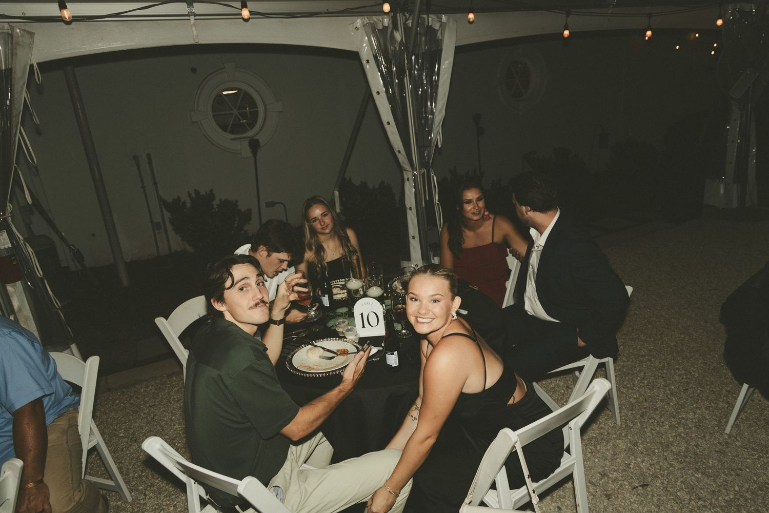 People gathered at an outdoor nighttime event sitting at round tables with white chairs under a dark tent.