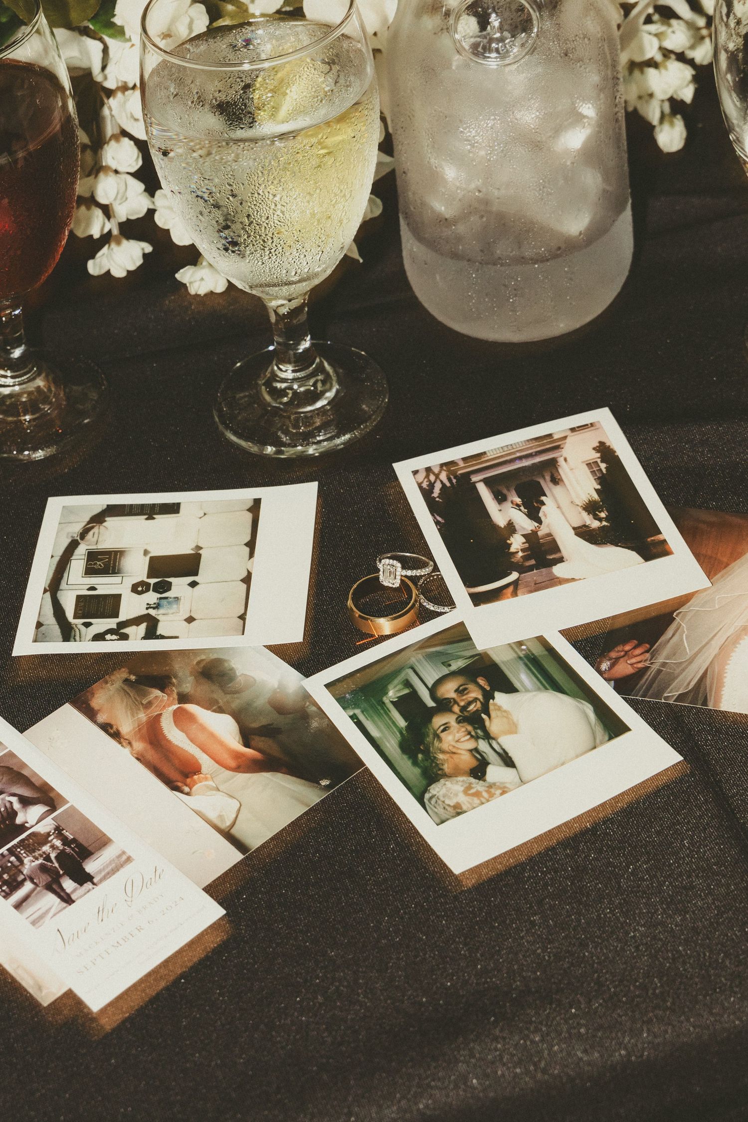 Vintage Polaroid photographs scattered on dark table next to wine glasses and white flowers create nostalgic ambiance.