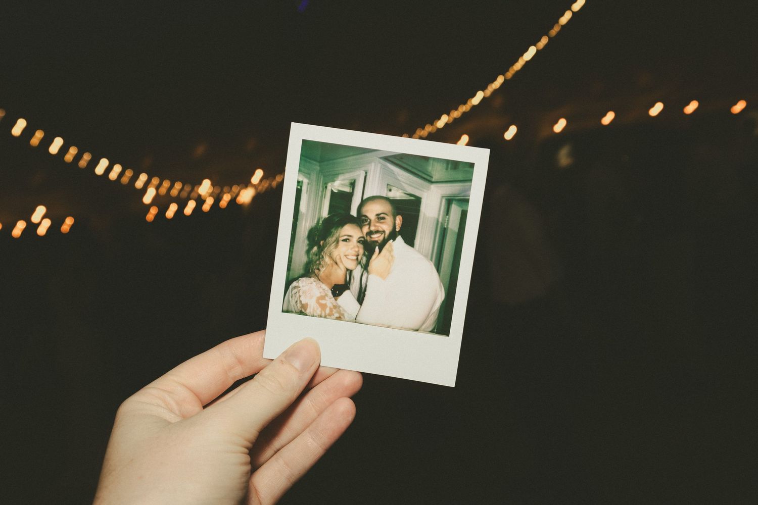 Hand holding a polaroid photograph against dark background with string lights illuminated in bokeh effect above.