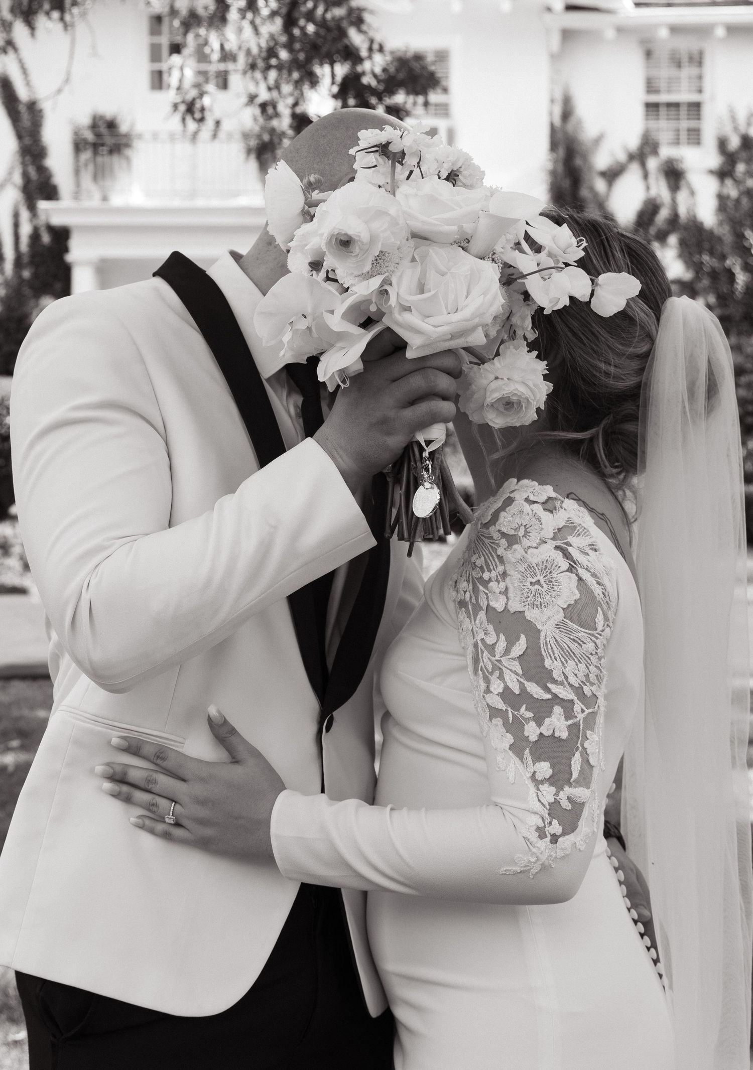 A romantic black and white wedding photo showing a tender kiss behind a white floral bouquet.
