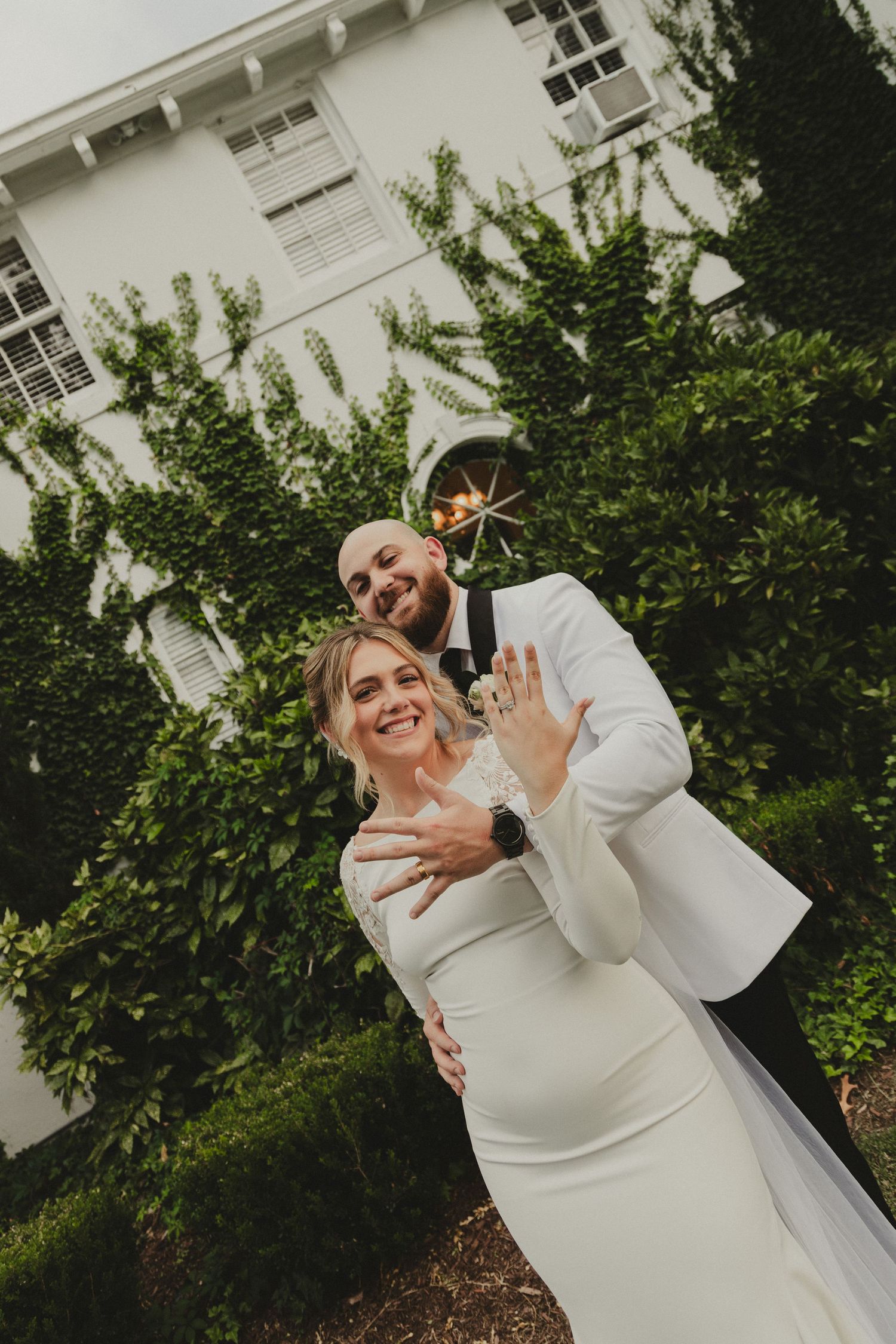 Newlyweds in white formal attire embrace and show off rings in front of a white house with green foliage.