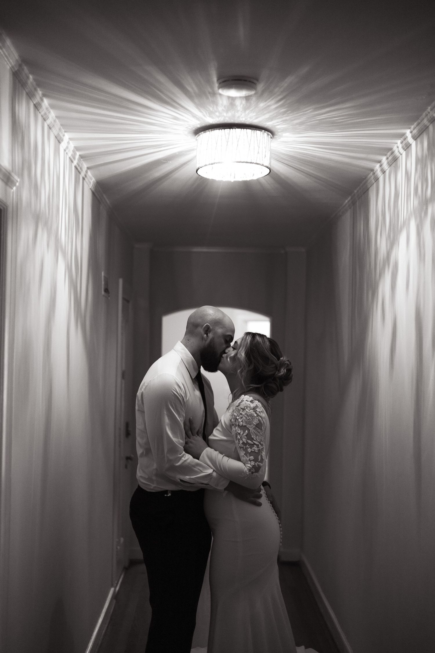 Romantic black and white photo of a couple kissing in a hotel hallway under dramatic ceiling light rays.