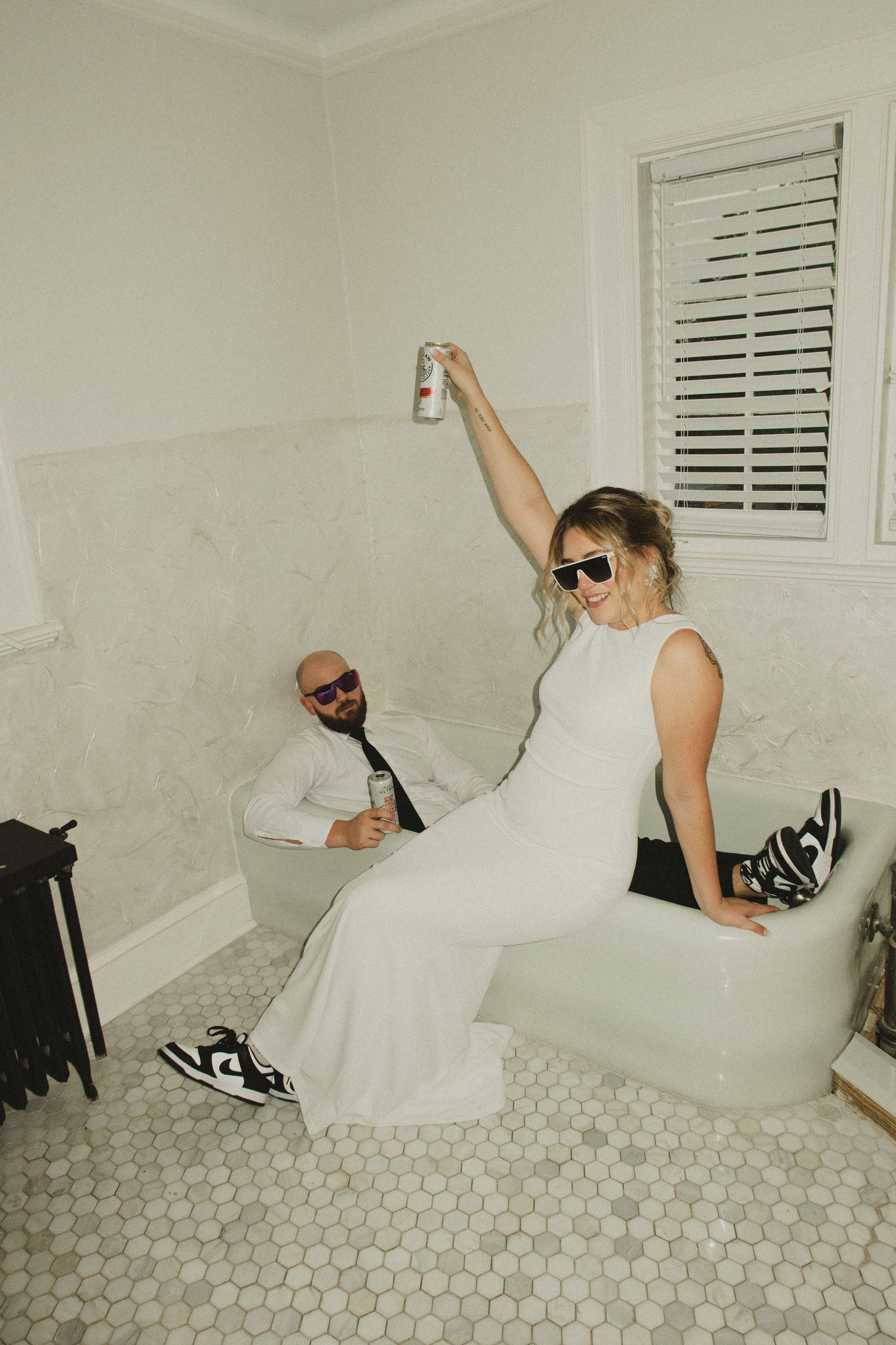 Fun moment captured in a bathroom with sunglasses and sneakers as two people pose casually on white tiled floor.