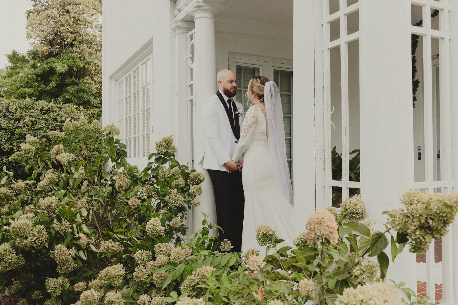 Two people stand together on an elegant white porch behind flowering hydrangea bushes.