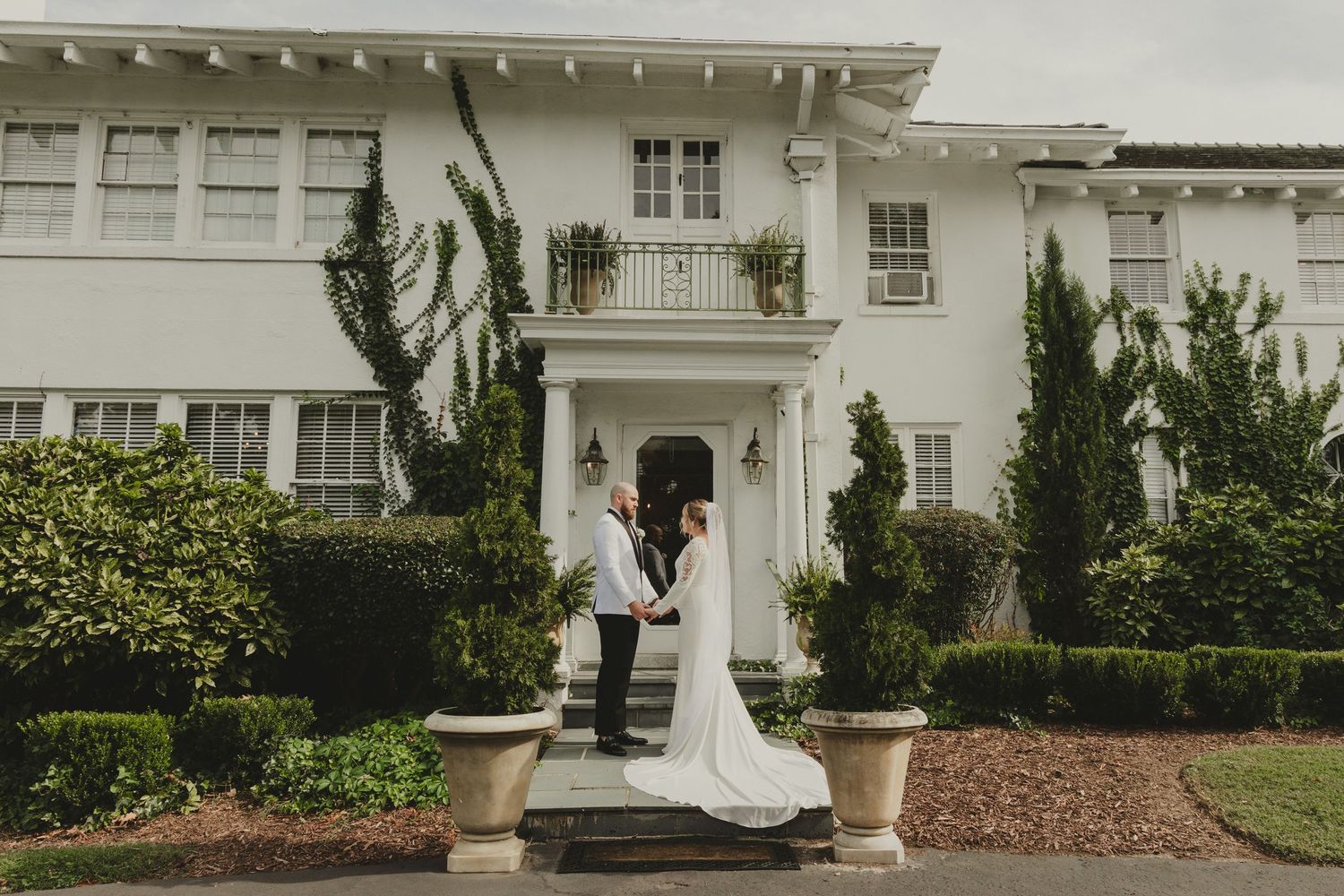 A romantic couple embracing on white steps in front of an elegant white mansion with landscaped gardens.