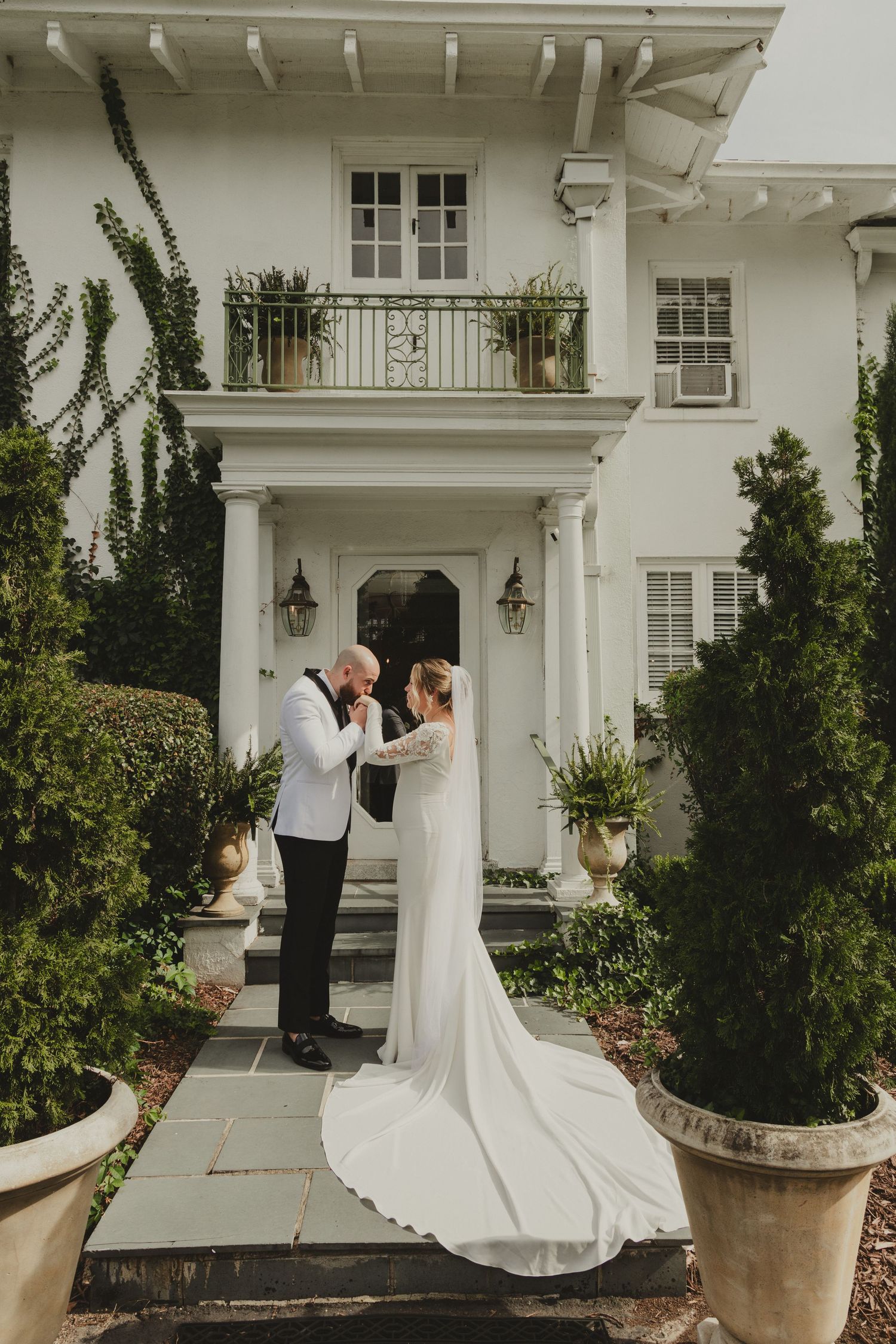 A romantic moment shared on the steps of an elegant white colonial mansion with lush greenery and trailing vines.