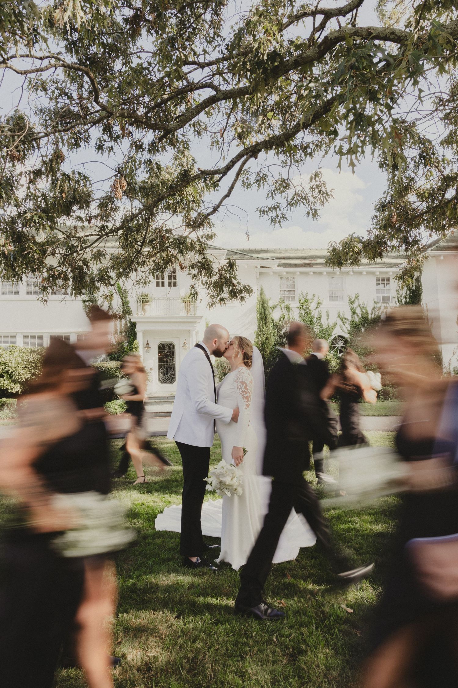 A romantic moment captured at an outdoor wedding ceremony with motion-blurred guests walking past under a large tree.