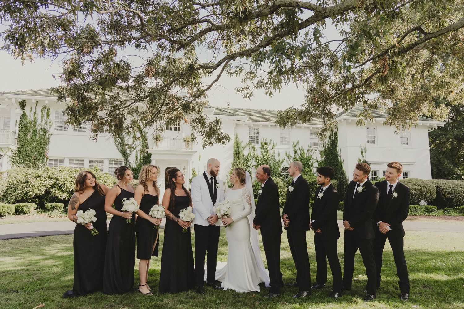 A wedding party poses together outdoors on a sunny day with bridesmaids in black dresses and groomsmen in dark suits.