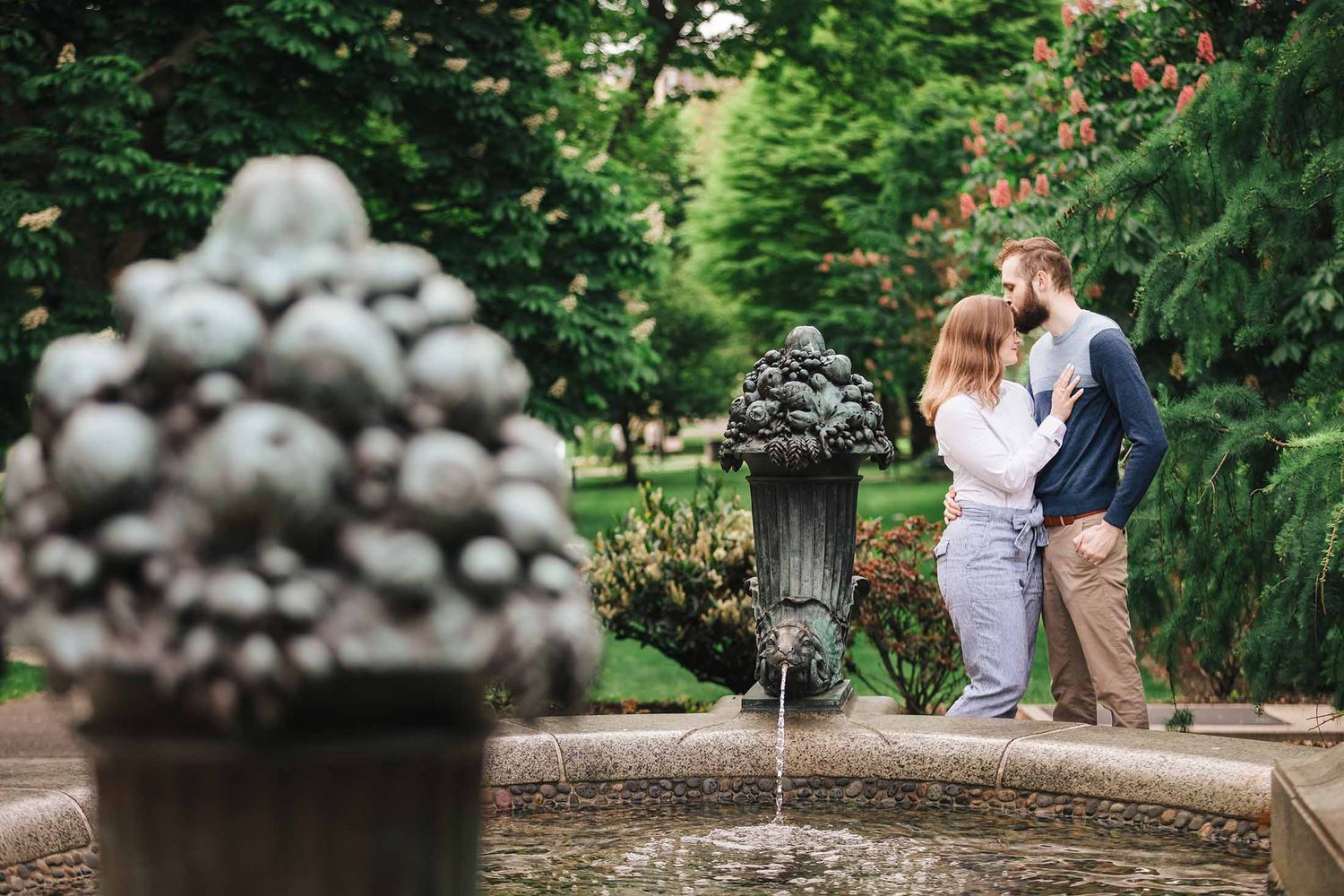 Groom-to-be gently kissing fiancée beside classical stone fountain at George Robert White Memorial during Boston engagement session
