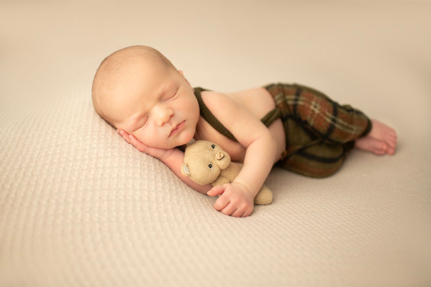 A sleeping newborn baby in plaid overalls cuddles with a small teddy bear on a cream-colored blanket.