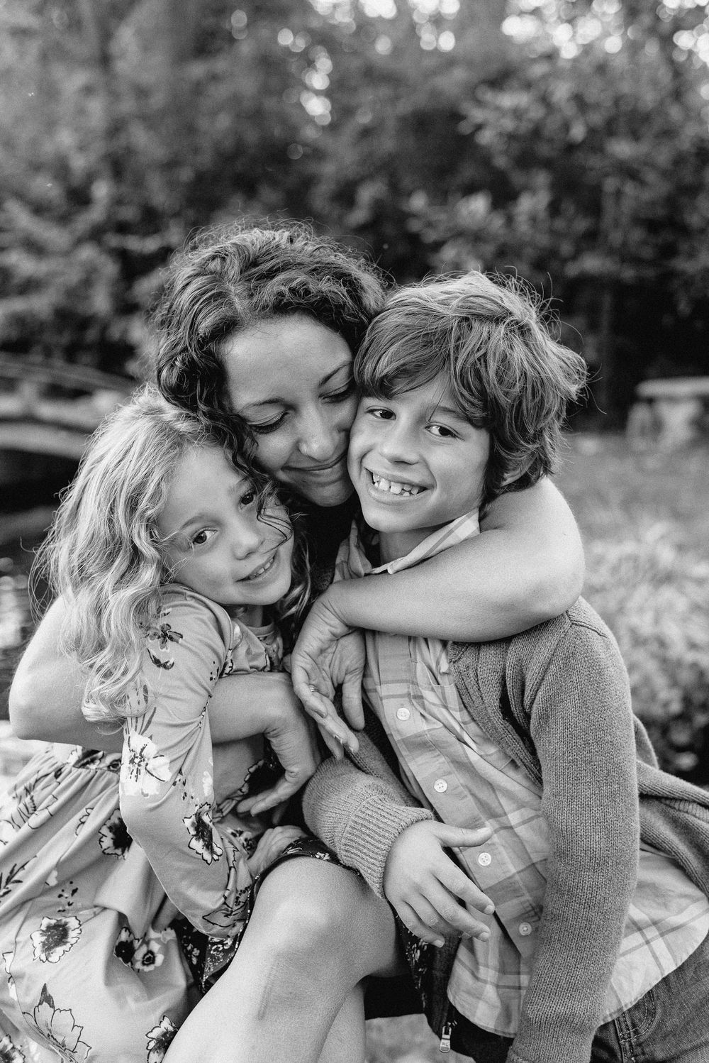 A black and white family portrait shows three people in a warm embrace outdoors with trees in the background.