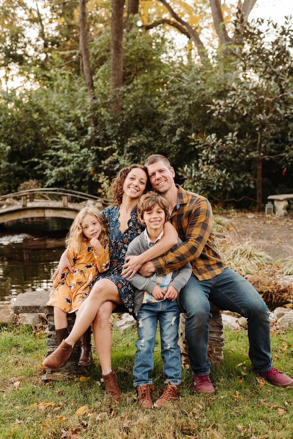 Family of four poses together on wooden bench near garden pond during fall photo session.