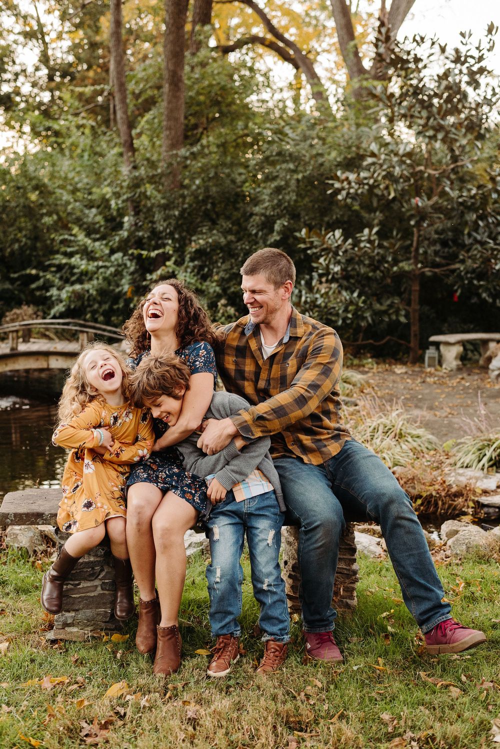 Family sits on rustic bench outdoors sharing genuine laughter during autumn photoshoot.
