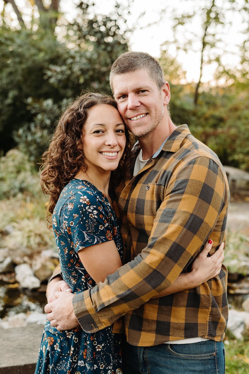 A couple embraces outdoors in autumn, with woman in floral dress and man wearing plaid flannel shirt.