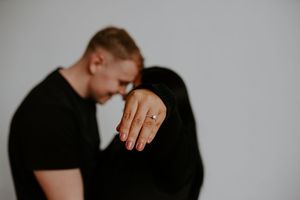A black and white studio portrait highlighting an engagement ring while a couple embraces.
