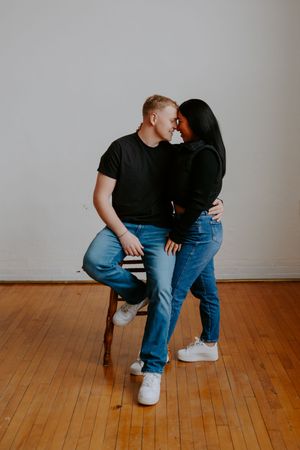 Couple in matching black tops and jeans share tender moment on wooden floor.