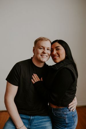 Couple in matching black tops and blue jeans embrace warmly against neutral background.