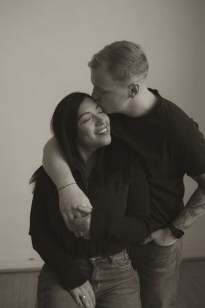 Intimate couple embraces while wearing black and denim against a neutral wall.