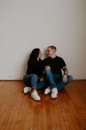 Casual couple shares playful moments while sitting against white wall.