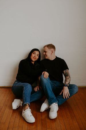 Couple in matching black tops and jeans share affectionate moment on wooden floor.