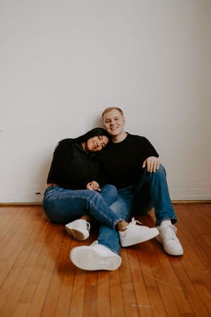 Couple lounging casually on wooden floor in casual attire against white wall.