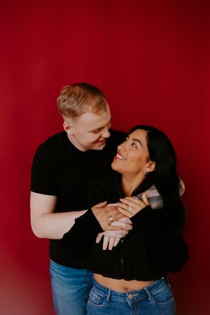 Romantic couple embracing against deep red backdrop in casual black and denim outfits.