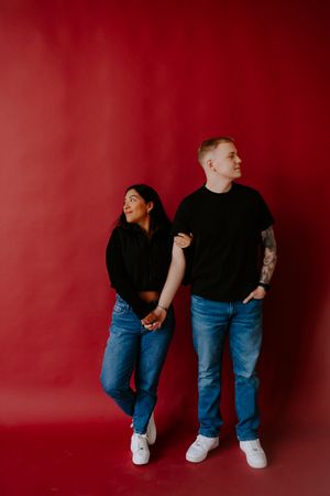 Couple in matching denim and black outfits posing playfully against red wall.