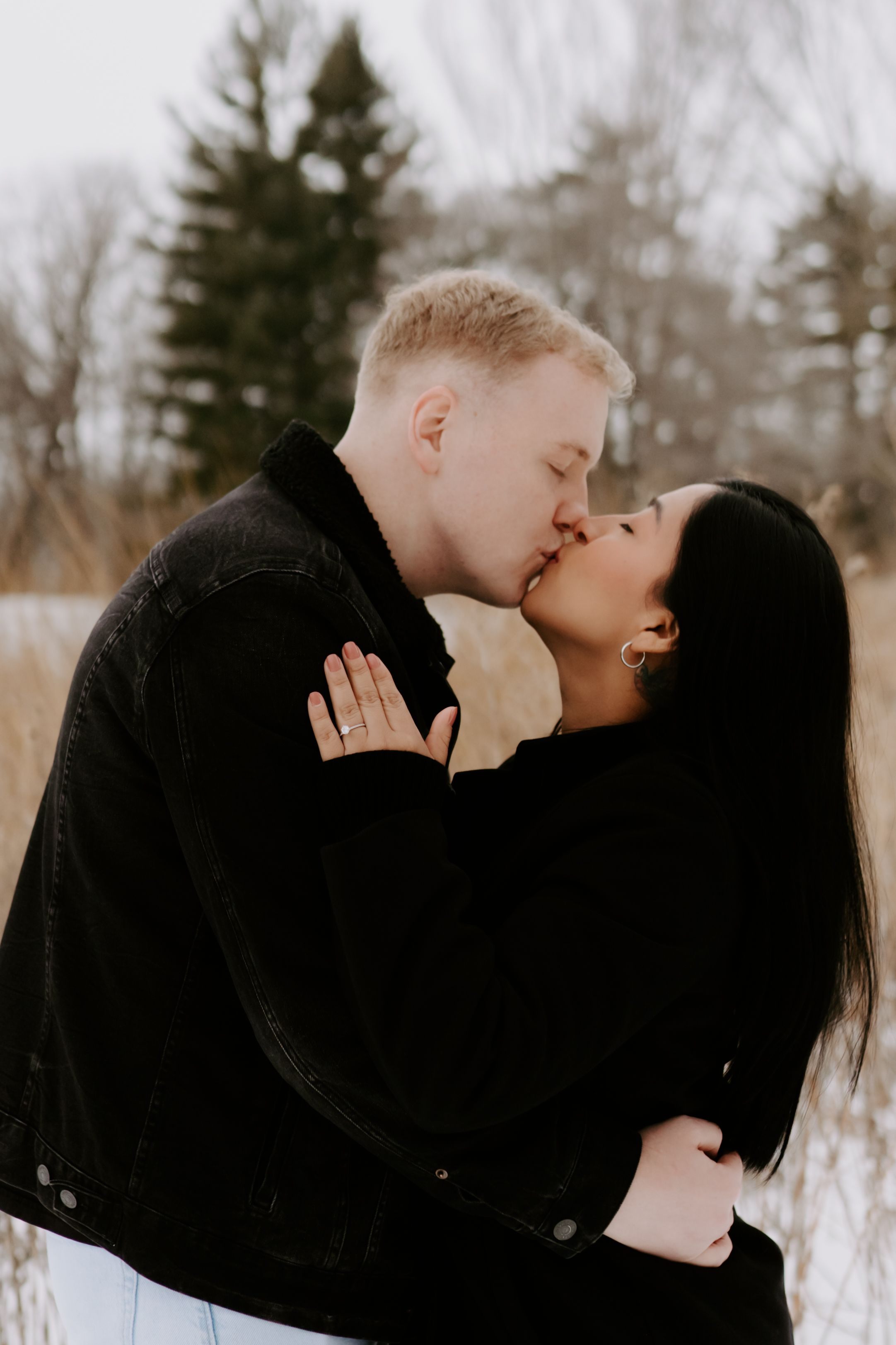A romantic winter photo shoot showing a couple embracing and kissing in the snow with evergreen trees in background.