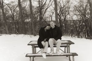 A couple sits together on a wooden bench in a snowy winter landscape with bare trees in the background.