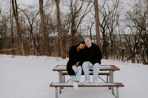 A couple sits close together on a wooden bench surrounded by snow and winter trees.