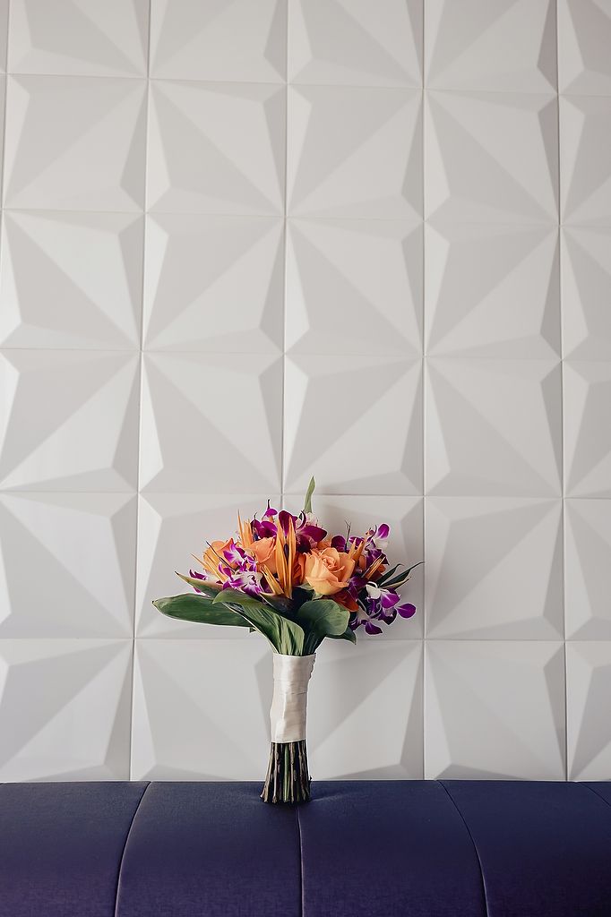 Vibrant bridal bouquet with orange lilies and purple accents sits on purple couch against geometric white wall background.