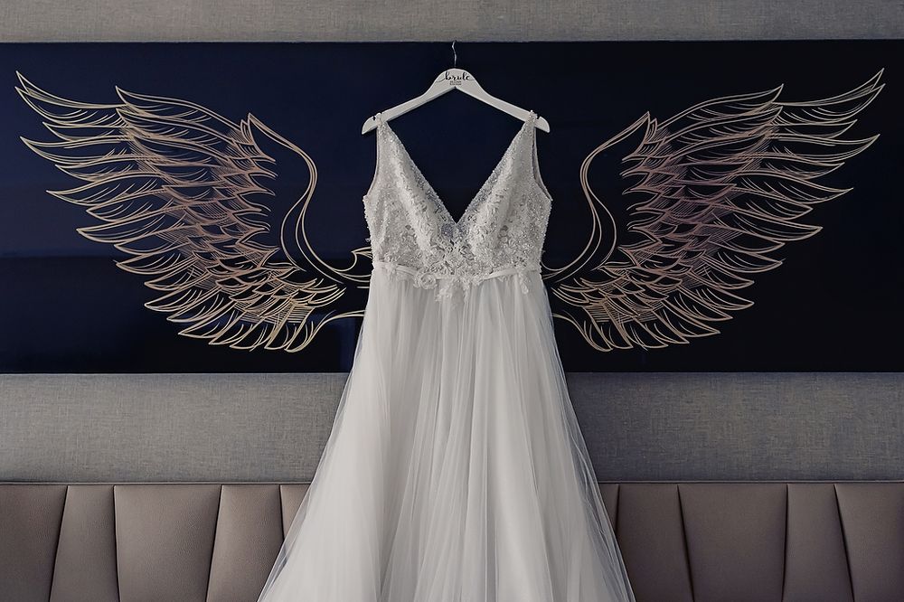 A white tulle wedding dress hangs centered between large metallic angel wings on a dark wall.