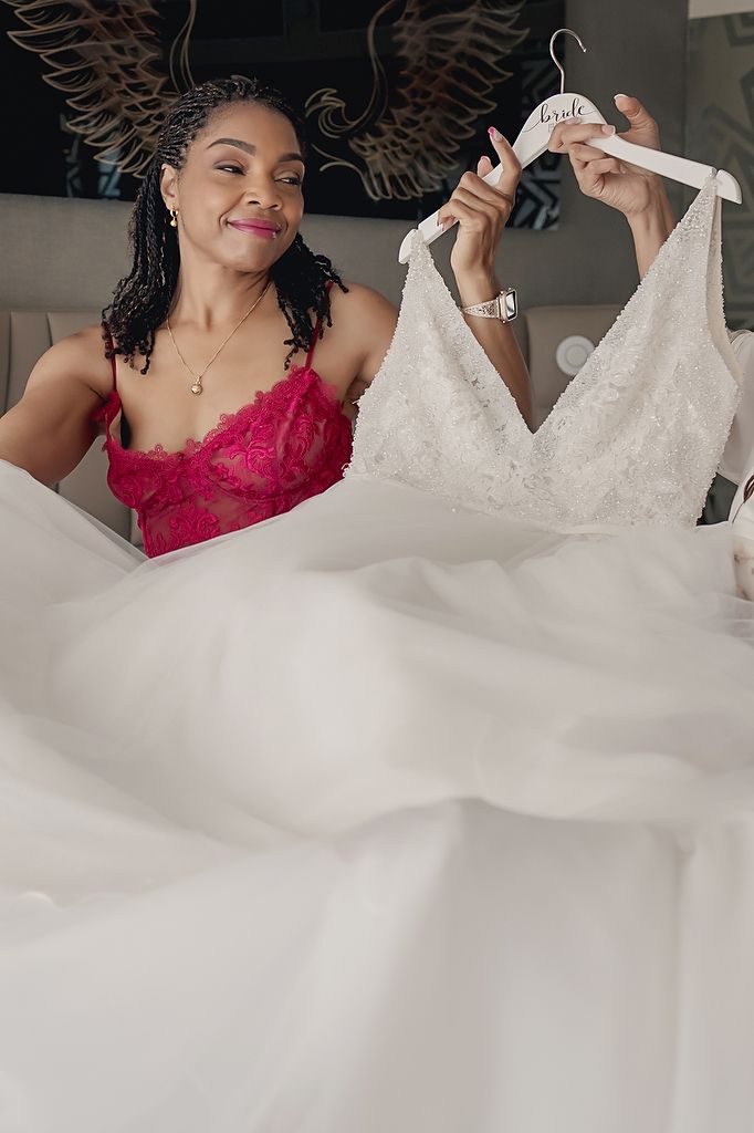 Someone wearing a red lace camisole admires and holds up a beautiful white wedding gown against a dark background.