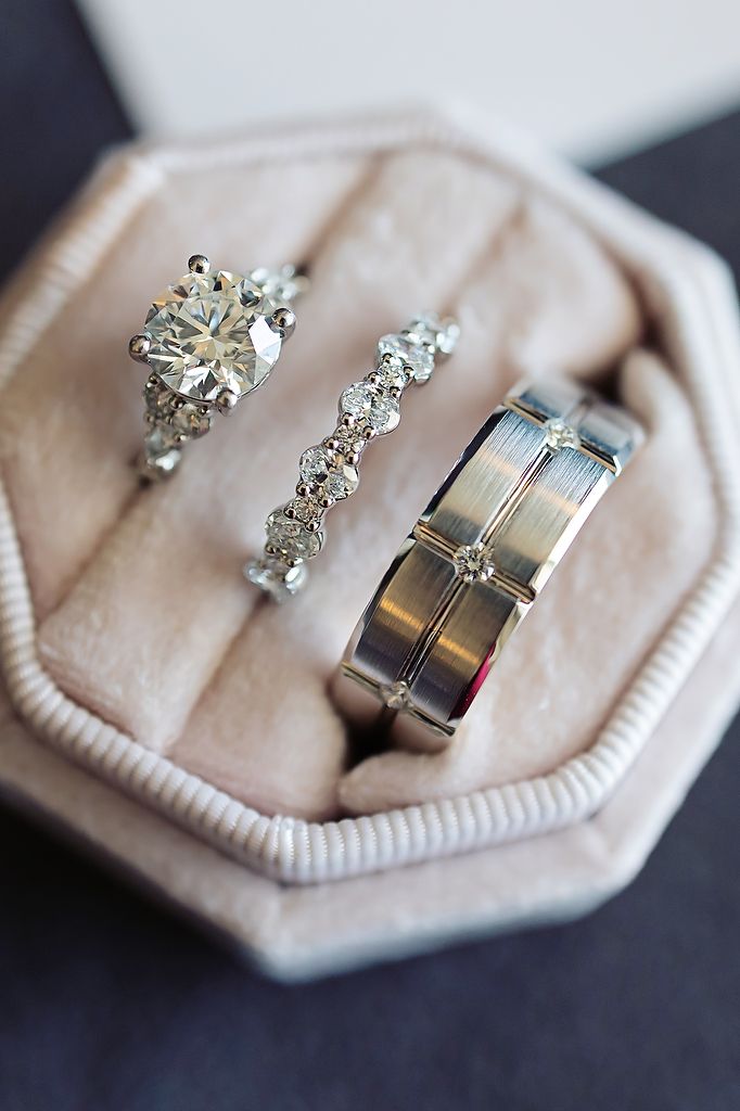 Three elegant wedding rings including a diamond solitaire, diamond band, and brushed metal men's band rest in a beige ring box.