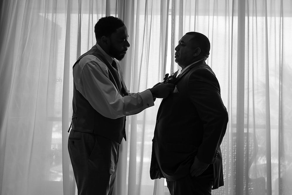 Two men in conversation stand in silhouette against sheer white curtains in a dramatic black and white scene.